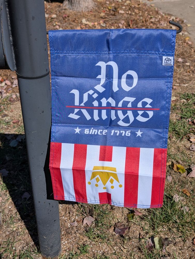 Photo of a garden flag hanging from a suburban mailbox post. The flag says No Kings since 1776 with an upside-down crown, printed over a blue banner at the top with vertical red stripes at the bottom. A small Flags for Good tag is visible in the upper corner.