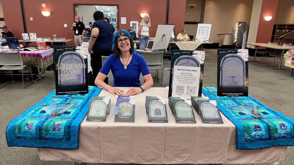 A woman with blue hair and a blue shirt sits at a table covered with a light brown tablecloth and two colorful blue quilted table runners. On the table are five stacks of books, and two stand up posters featuring covers of her Ghostly Tales series.