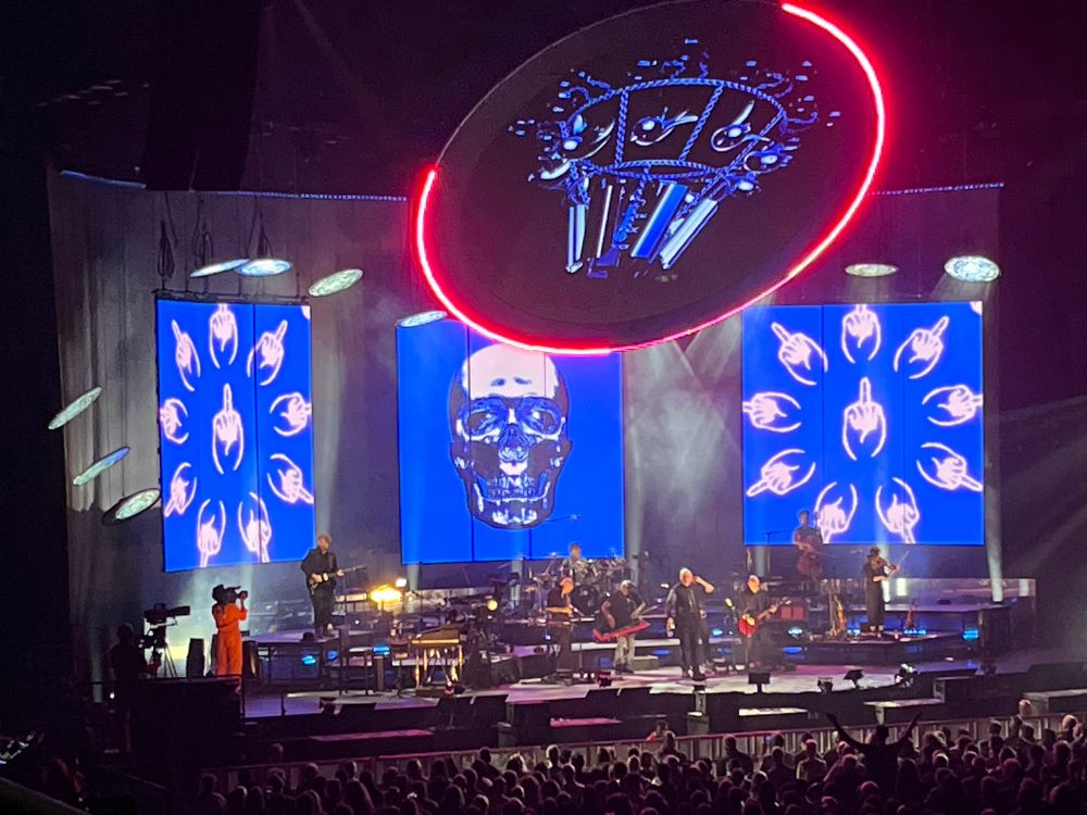 Peter Gabriel and his band on a stage in front of the giant screen with art featuring hands displaying the middle finger and a skull on a blue background.