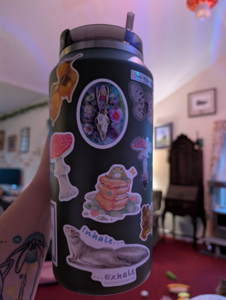 A green 64 oz Stanley mug with a number of stickers on it. There's a frog on a stack of pancakes, a squirrel splooting captioned "inhale...exhale", nore mushrooms, a moth, a deer skull with flowers...