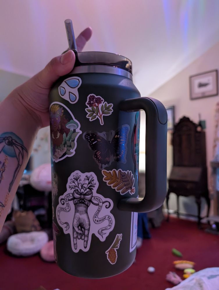 A green 64 oz Stanley mug with a number of stickers on it. The other side of the Peculiarity Shop sticker, a water drop sticker, a fern sticker, a pine cone, another moth, and a black and white hand with herbs around it that has a ribbon that says NO JUSTICE, NO PEACE / KNOW JUSTICE, KNOW PEACE
