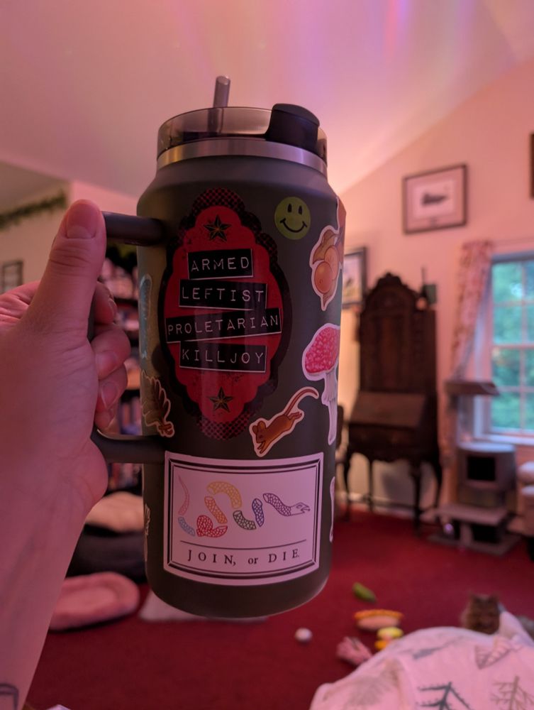 A green 64 oz Stanley mug with a number of stickers on it. There is one that is red and says ARMED LEFTIST PROLETARIAN KILLJOY, and another with a rainbow snake split into sections that says JOIN OR DIE. There is also one of those '90s 🙂 stickers that are scratch and sniff and a little mouse and a mushroom.