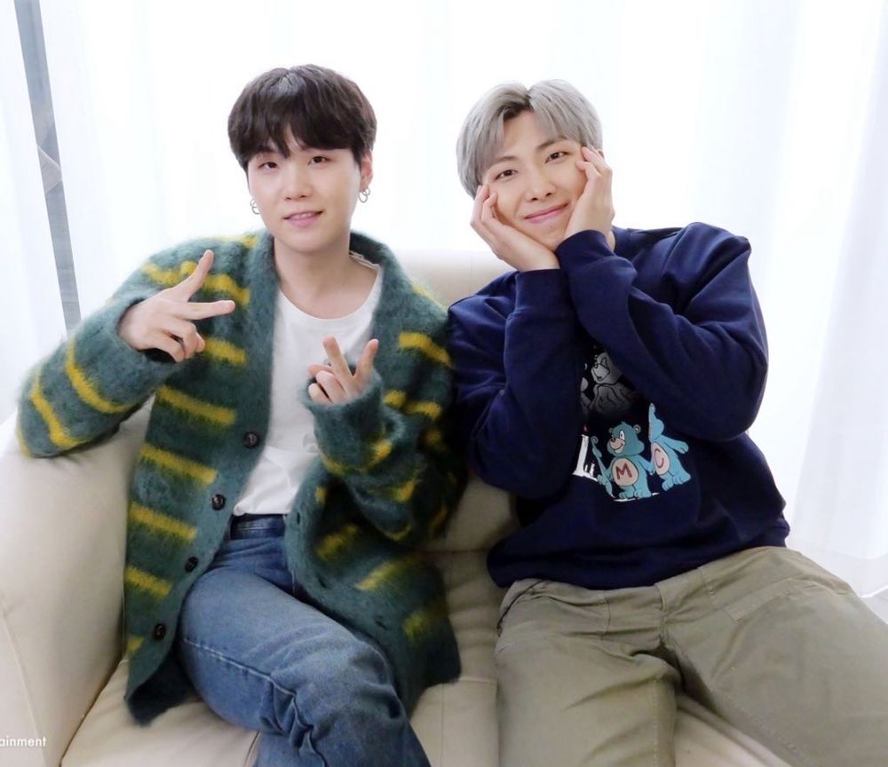Yoongi and Namjoon sitting next to one another. Yoongi is wearing blue jeans, a white tshirt and a fuzzy, unbuttoned green cardigan with yellow stripes and big brown buttons. His hair is black and he’s holding his hands up and giving peace signs with  both hands, the back of his hands facing the camera. Namjoon is wearing a navy blue sweater with light blue carebears on it and khaki pants. His hands are cupping his own cheeks. His hair is light grey. 
