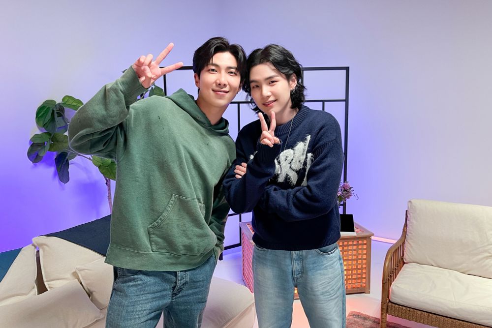 Namjoon & Yoongi on the set of the first episode of Suchwita when Namjoon was Yoongi’s guest. They are standing side by side leaning into one another with their heads almost touching. They both have black hair, Yoongi’s is long and soft waves frame his face and neck. He’s giving a peace sign with his left hand and wearing a navy blue sweater with white text on the front and light wash jeans. Namjoon is also doing a peace sign with his left hand and he’s wearing an oversized sea green hoodie and blue jeans. 