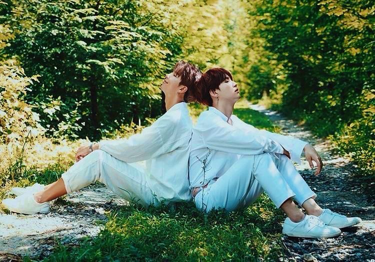 Yoongi and Namjoon sitting on the ground in a forest both wearing long sleeved white shirts, white pants, and white sneakers with no socks. They are sitting back to back with their heads tipped back againstt one another. Their eyes are closed. Namjoon is smiling and Yoongi’s face is calm. Namjoon’s hair is sandy brown and yoongi’s is dark reddish brown 