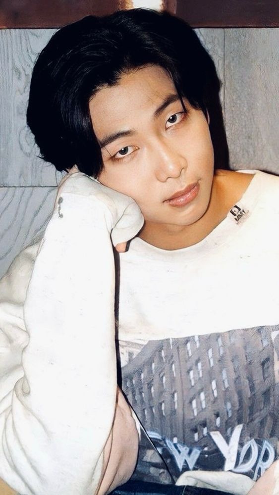 Kim Namjoon from November 2023. He’s in a long sleeved cream colored shirt that has been distressed at the collar and sleeves and has a screen print of buildings and the words “NEW YORK” on the front. He’s looking at the camera, his head is tilted and he’s resting his cheekbone on his fist. His hair is long and black. 