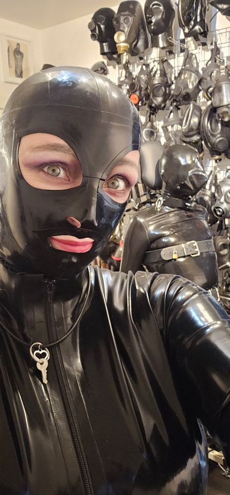 Black Latex covered women with a wall of gasmasks behind her she is wearing a chasity key