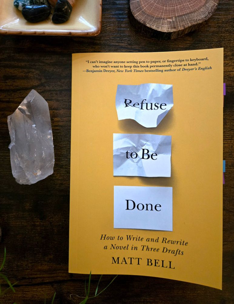 A book, "Refuse to be Done" by Matt Bell, on a brown wooden surface. Around it are a wooden coaster, dish with polished stones, and a quartz crystal. 
