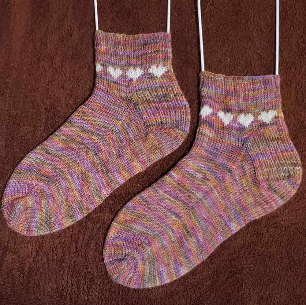 two hand-knit ankle socks are pastel rainbow speckles with white hearts around the ankle, and four hearts are showing on each. The socks are on white metal sock blockers. The background is a chocolate brown terrycloth towel.