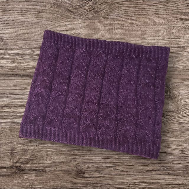 rich deep purple lacy work cowl. The lacy pattern is repeated for 7 rows creating soft peaked points with 1x1 ribbing on top and bottom. the luxurious suri silk held with hemlock sock yarn makes for a plush fabric. The garment is displayed on a lightly faded, medium-toned wooden background.