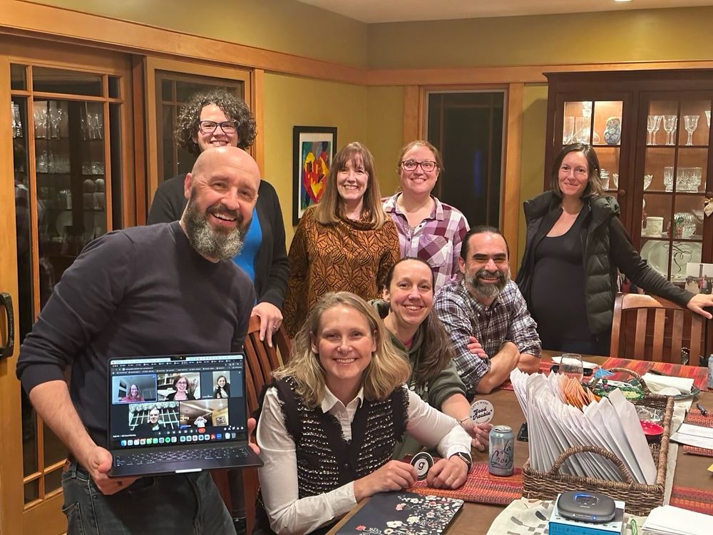8 people gathered around a table (3 sirring, 5 standing). Man with beard in foreground (left corner) holding a computer displaying 5 squares on a Google Meet
