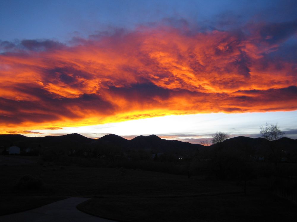 Image of dramatic, fiery orange and yellow clouds in a darkening blue sky, stretching over a black silhouette of mountains to form a breathtaking sunset scene. 