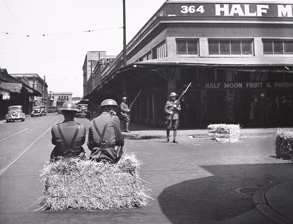Black and white image with two national guard soldiers sitting on a hay bale in the foreground, two more soldiers with bayonets in the mid ground, and a building with a produce stand on the right side. 