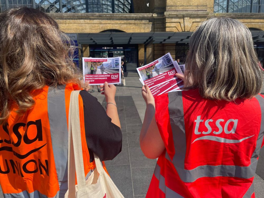 Two women with their backs to us, facing Kings Cross station. They are wearing TSSA-branded Hi-vis vests and holding leaflets for TSSA's campaign for better funding for British Transport Police.