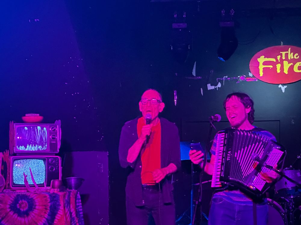 Two men singing on a stage at a venue called The Fire. I'm the one holding an accordion. There are analogue TVs just sitting to the side showing random patterns and static.