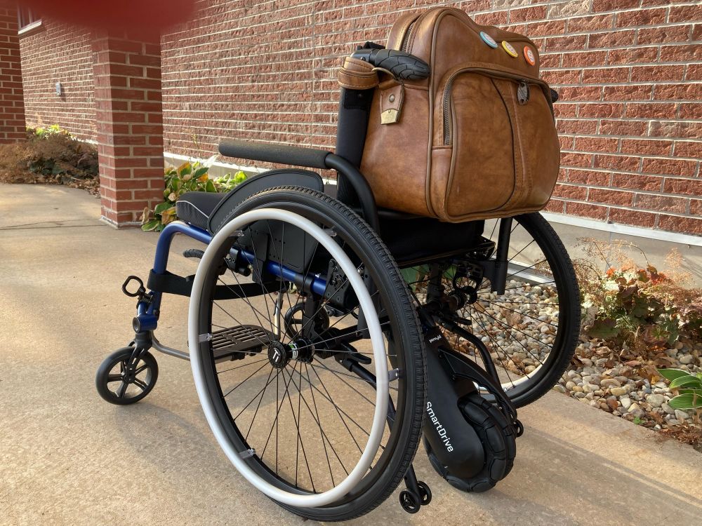 the same wheelchair seen from behind, giving a better view of the power assist drive. there’s also more of the background, revealing it’s a cover walkway with brick pillars next to the brick exterior of a building. the wheelchair is parked alongside the flowers by the wall