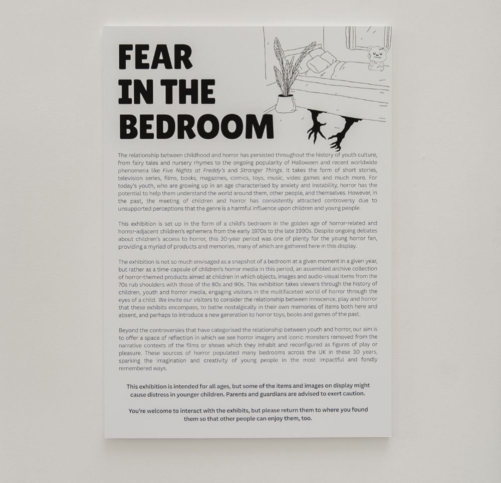 Exhibition wall showing title: FEAR IN THE BEDROOM. B+W illustration shows a bed with a zombie teddy on top and monster hands stretching out from underneath. Text below reads:
"The relationship between childhood and horror has persisted throughout the history of youth culture, from fairy tales and nursery rhymes to the ongoing popularity of Halloween and recent worldwide phenomena like Five Nights at Freddy’s and Stranger Things. It takes the form of short stories, television series, films, books, magazines, comics, toys, music, video games and much more. For today’s youth, who are growing up in an age characterised by anxiety and instability, horror has the potential to help them understand the world around them, other people, and themselves. However, in the past, the meeting of children and horror has consistently attracted controversy due to unsupported perceptions that the genre is a harmful influence upon children and young people.  
 
This exhibition is set up in the form of a child’s bedroom in the golden age of horror-related and horror-adjacent children’s ephemera from the early 1970s to the late 1990s. Despite ongoing debates about children’s access to horror, this 30-year period was one of plenty for the young horror fan, providing a myriad of products and memories, many of which are gathered here in this display.  
 
The exhibition is not so much envisaged as a snapshot of a bedroom at a given moment in a given year, but rather as a time-capsule of children’s horror media in this period, an assembled archive collection of horror-themed products aimed at children in which objects, images and audio-visual items from the 70s rub shoulders with those of the 80s and 90s. This exhibition takes viewers through the history of children, youth and horror media, engaging visitors in the multifaceted world of horror through the eyes of a child. [continued in next image]