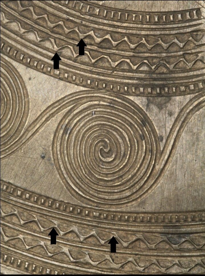 A bronze plate decorated with an inserted pattern of spirals, triangles and lines. The decoration is inserted using a bronze chisel in a bronze sheet. The individual tool marks are visible at highlighted with black arrows. 