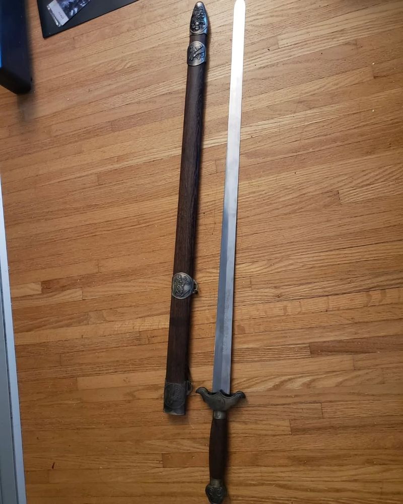 a decorative sword with a wooden and bronze handle, unsheathed next to its case, which is made up of wood with bronze details. the sword and case are laid out flat on a wooden floor