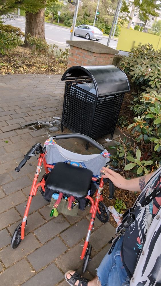 A photo of a red and black rollator style walker in front of a bush and trash can. A person with tattoos leans on the handle, joint in hand, but is mostly cropped from the image. The walker has a diy bag hanging off the front that partially shows the anime character Chopper, who is a cute reindeer in a hat, nd is covered in stickers and pins, beaded Kandi bracelets hanging off the bar. Under the seat hangs another, smaller diy bag