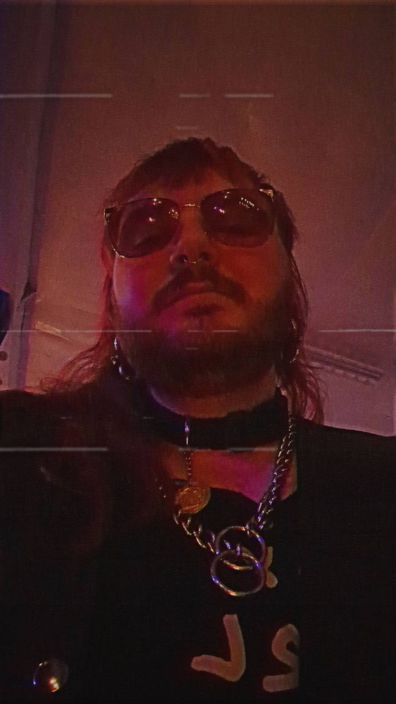 selfie in red lighting of a bearded nonbinary punk with a red mullet. they have a gold septum ring, and silver ear weights that look like snakes. they are wearing aviators with red tinted lenses, a black dog collar, and a silver chain. their outfit is mostly black, but you can see a bit of light text on the shirt