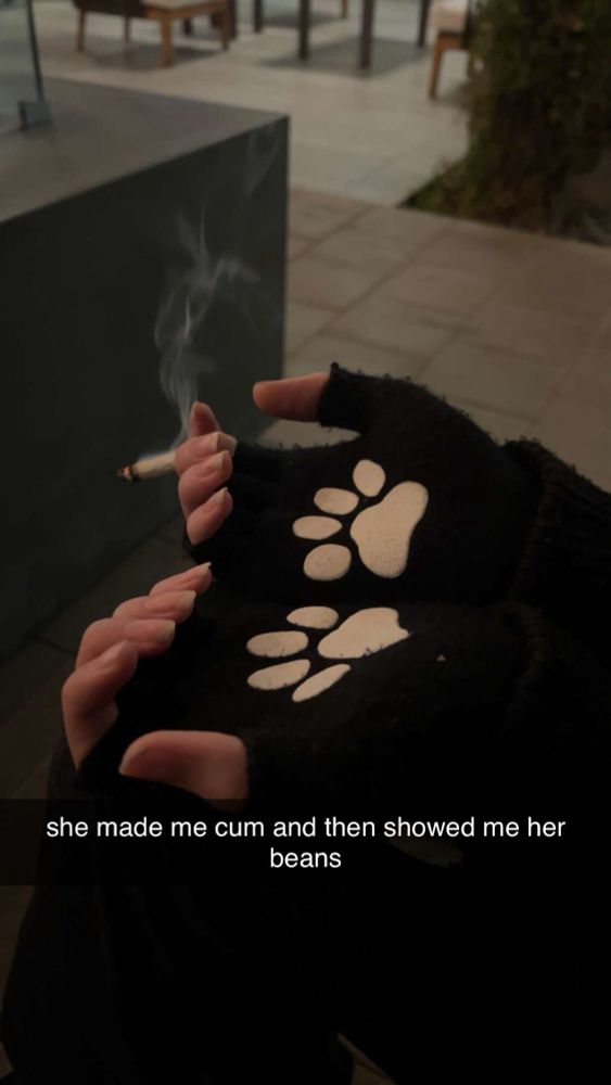 a screenshot of a snapchat image if someone smoking a cigarette looking joint outdoors while wearing fingerless black gloves with white puppy paw bean patterns on them and a caption that says “she made me cum and then showed me her beans”