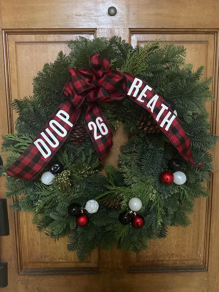 A Christmas wreath with decorative ball ornaments and a black and red checkered ribbon with DUOP REATH 26 on it