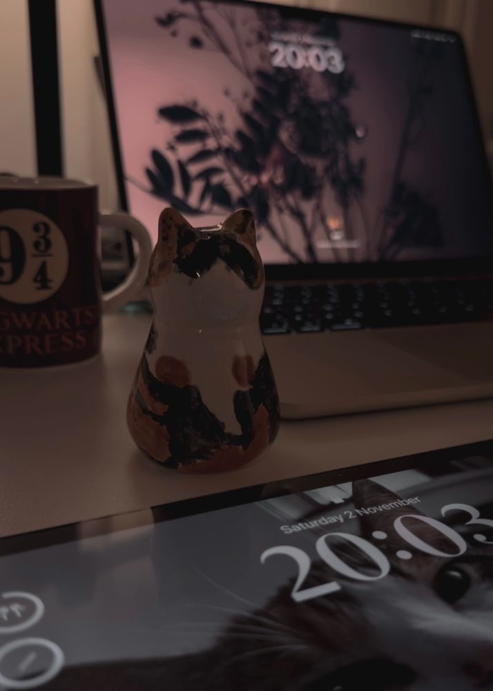 Painted figure of a calico cat on a desk in front of a laptop and iPad, next to a 9 3/4 Hogwarts Express mug. 