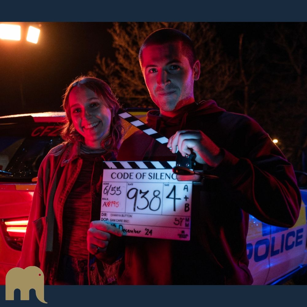 A square image of two people standing outside in front of a police car at night. On the left is Rose Ayling-Ellis her hair is down, she is smiling at the camera. She is dressed in a casual top, jeans and denim style jacket. On the right is Kieron Moore; his hair is in a short buzz cut and he is also smiling at the camera. He is wearing a dark hoodie and holding the clapperboard for Code of Silence.