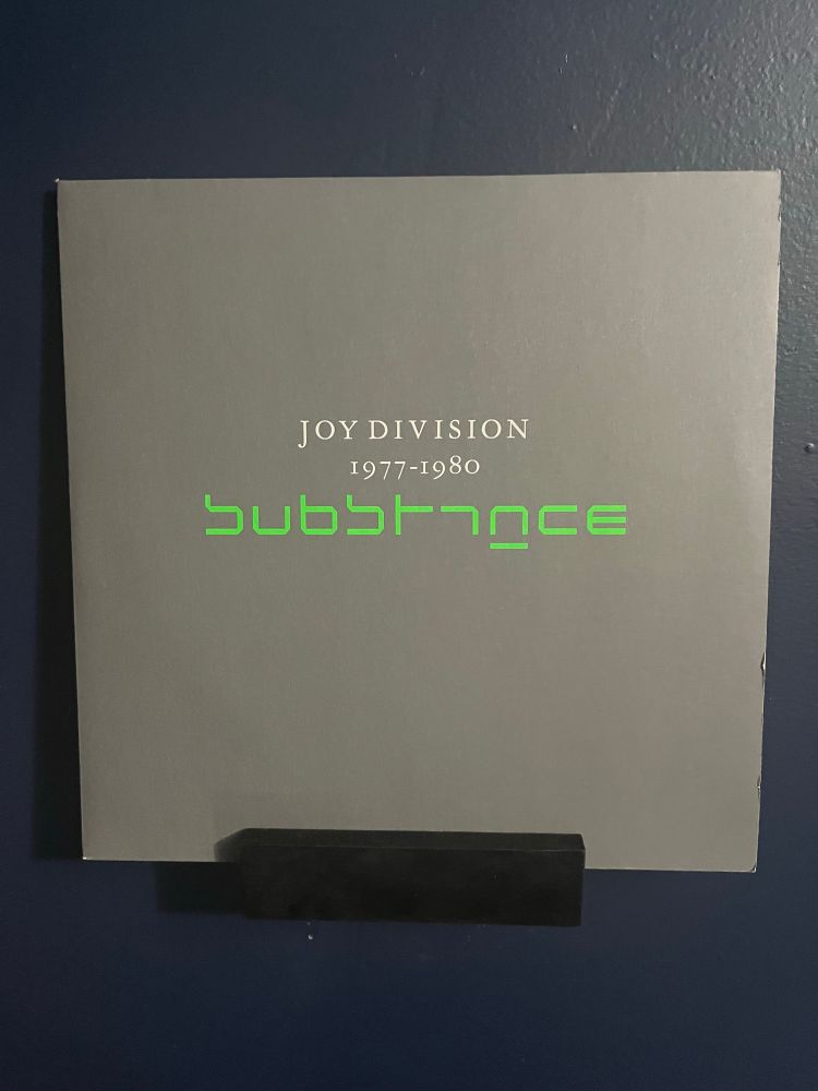 Album cover for the Joy Division compilation album, Substance 1977-1980. The cover is gray with green text saying the album title and band name. The album is displayed on a small black shelf on a dark blue wall. 
