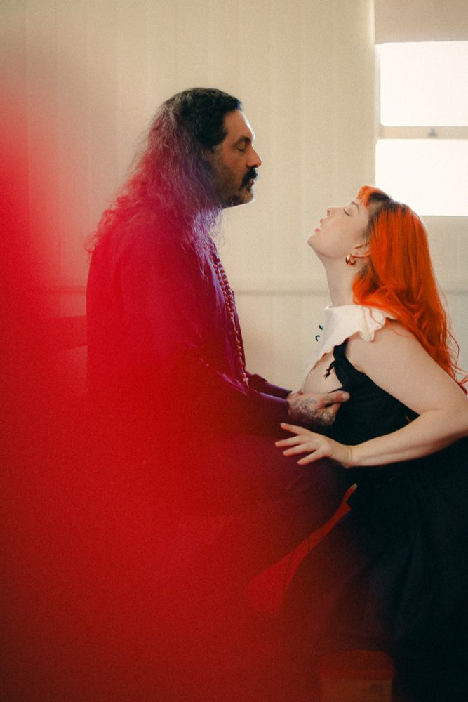 Redhead model wearing nun garb, seductiveky kneeling at the foot of a male priest figure as he opens her corset. Photography by Phthonos. 