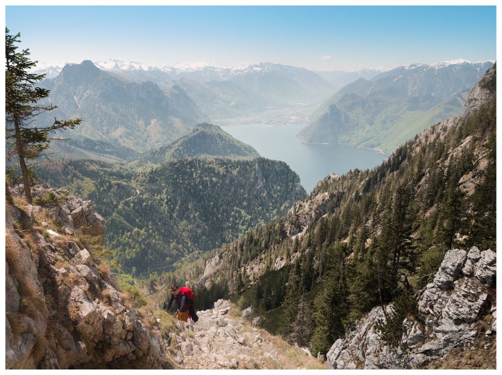 A hiker with a red backpack descends a rocky mountain path, overlooking a vast alpine landscape with a lake nestled between forested mountains and snow-capped peaks in the distance, likely the view from or of Traunstein in Austria.