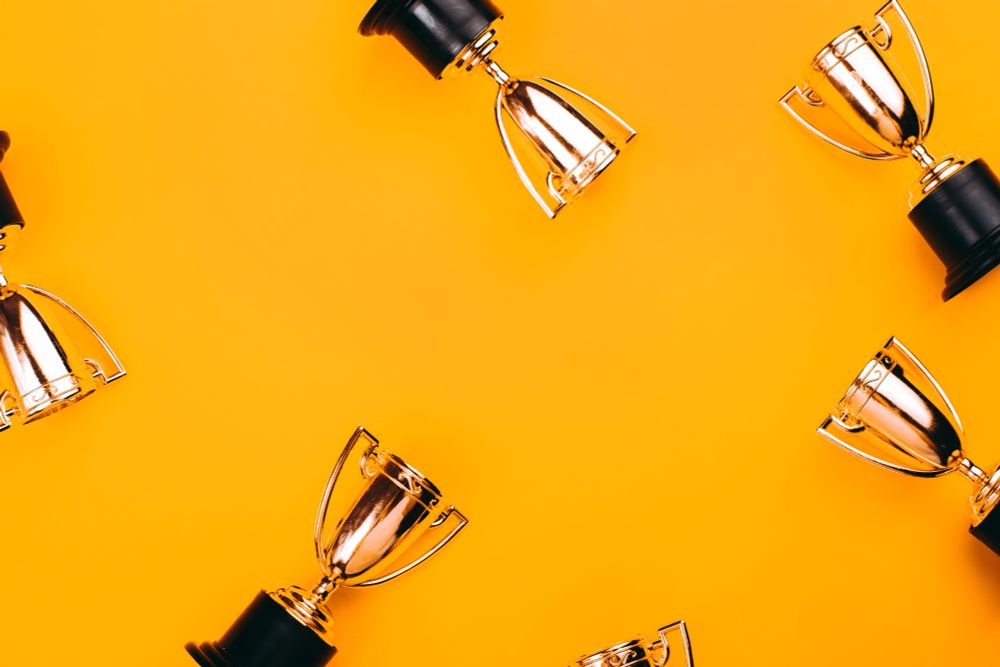 A flat lay image of multiple small gold trophies with black bases arranged around the edges of a vibrant orange background, leaving space in the centre. The image evokes celebration and achievement.