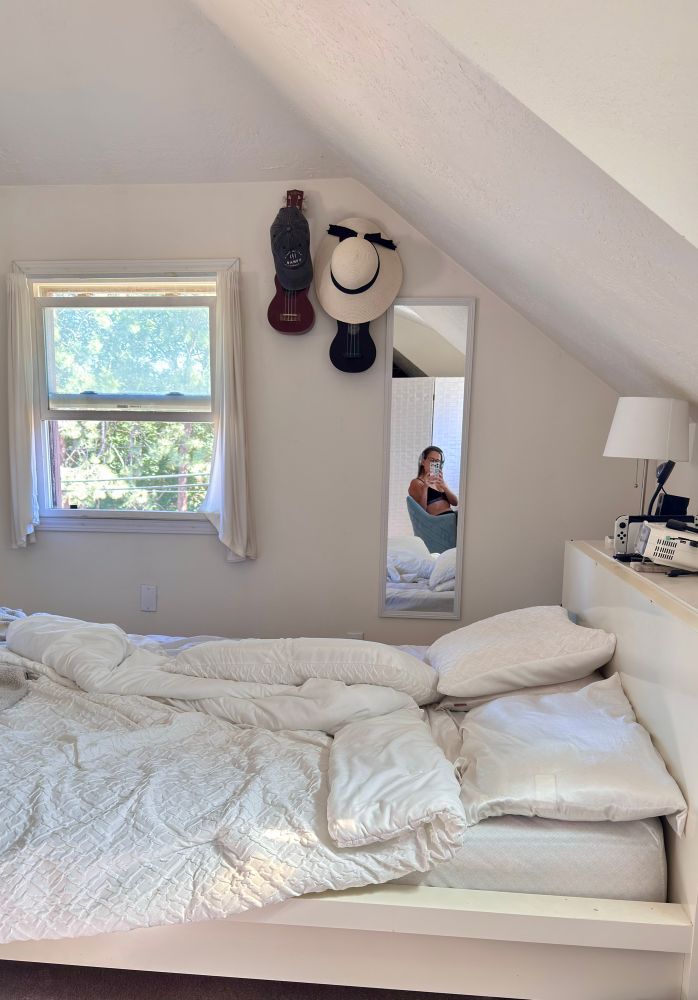 Messy bed with all white bedding and mirror selfie of Rachel in the background 