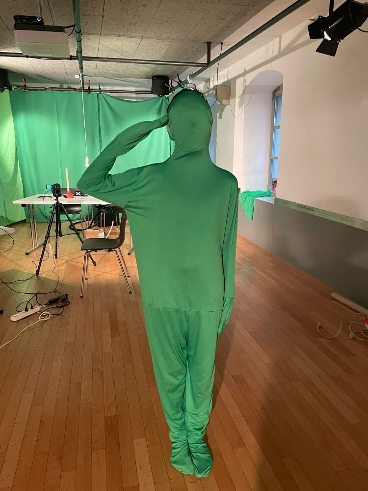 Eine Person in einem grünen Ganzkörperanzug steht salutierend da. Im Hintergrund ist eine Kamera auf einem Stativ und ein Teil eines Selfmade-Greenscreen zu erkennen.