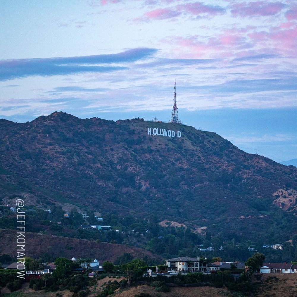 View of the Hollywood Hills at dusk, featuring the iconic Hollywood sign. A tall communication tower rises beside the sign, surrounded by rolling hills and scattered homes in the foreground. The sky above is painted with soft pink and blue hues.
