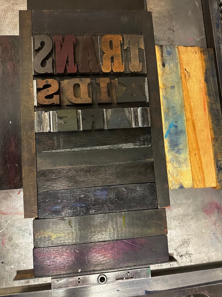 Wood type in the bed of a press reading (backwards) “Trans Kids Are [Blank]”