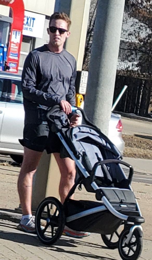 A male jogger stands at an intersection waiting for a light. He is pushing a running style baby stroller with nothing in it.