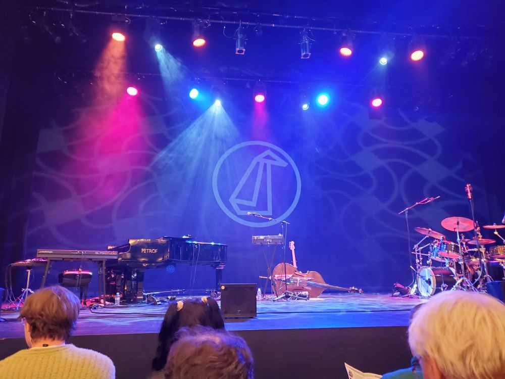 An empty stage showing a piano, a double bass, a drum kit. The stage is lit, with the logo of a circle and an abstract penguin. No performers are on stage yet. Crowd waits in anticipation 
