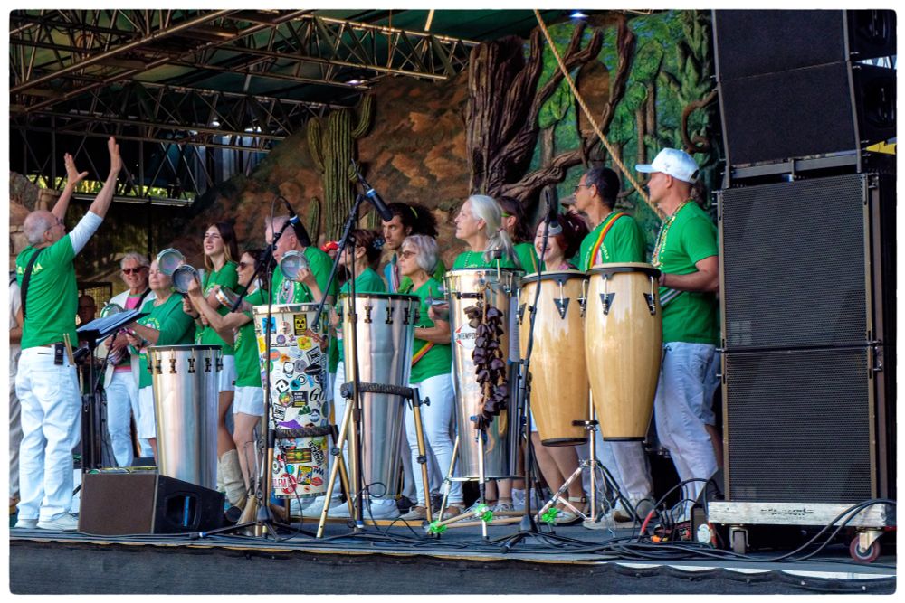Portland samba band Bloco Alegria performing on the zoo stage. A row of tall drums stands in the front, and at least a dozen musicians wearing green shirts and white pants stand in a row behind them. On the right is a bank of speakers, and on the left a man conducting the band stands with his arms raised and his back to the audience.