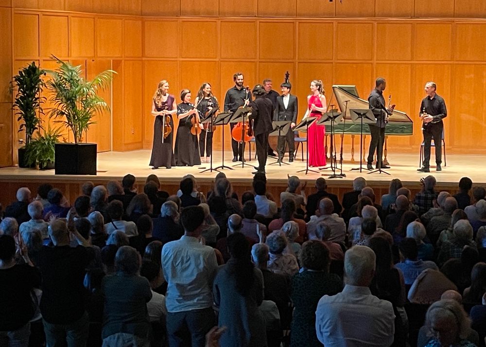 Musicians gathered on a stage with classical instruments including a harpsichord 