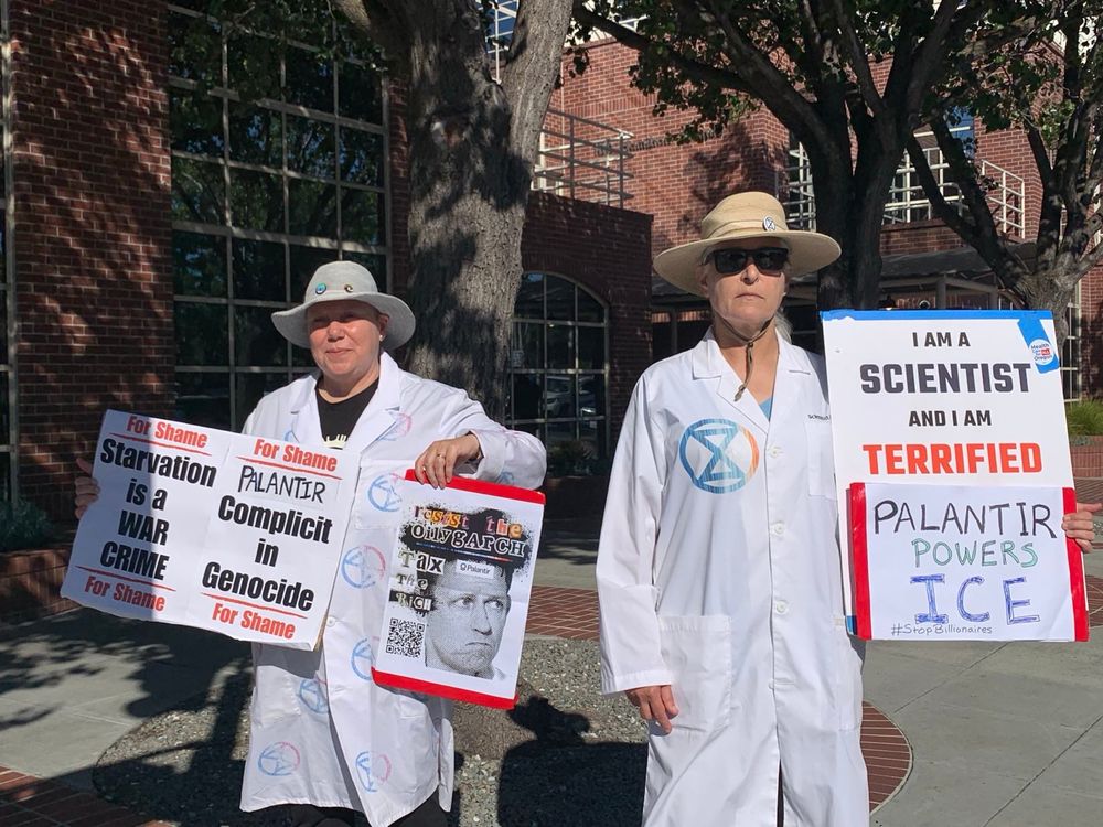 Two people standing in a plaza with trees and a brick and glass building in the background housing the company Palantir. They are wearing labcoats emblazoned with the Scientist Rebellion logo and holding signs saying "Starvation is a WAR CRIME", "Palantir complicit in genocide", "I am a scientist and I am terrified", and "Palantir powers ICE". Another poster has a graphic of Palantir founder Peter Theil with "Resist the Oilygarch, Tax the Rich". 