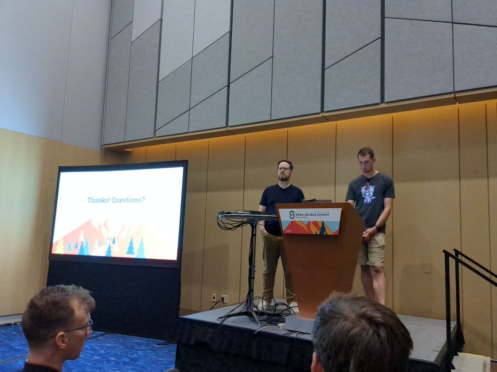 Two men stand at a podium delivering a conference talk with a projector screen in the background