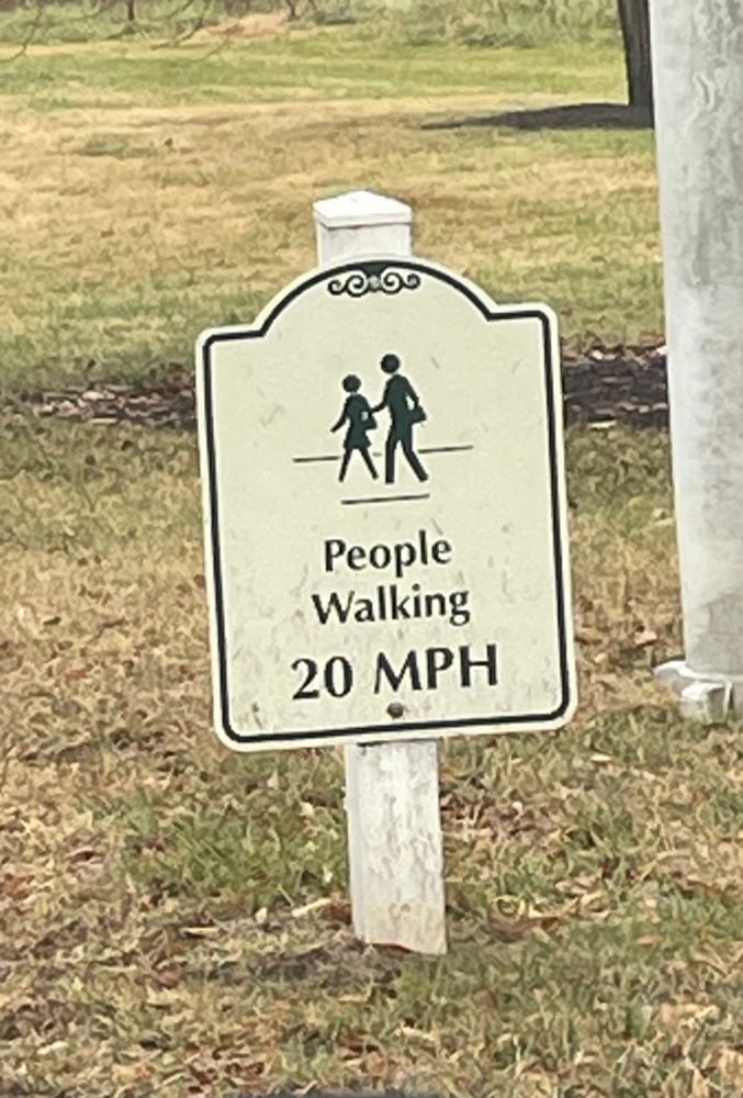 Small sign in grass beside roadway reads “people walking 20 mph”
