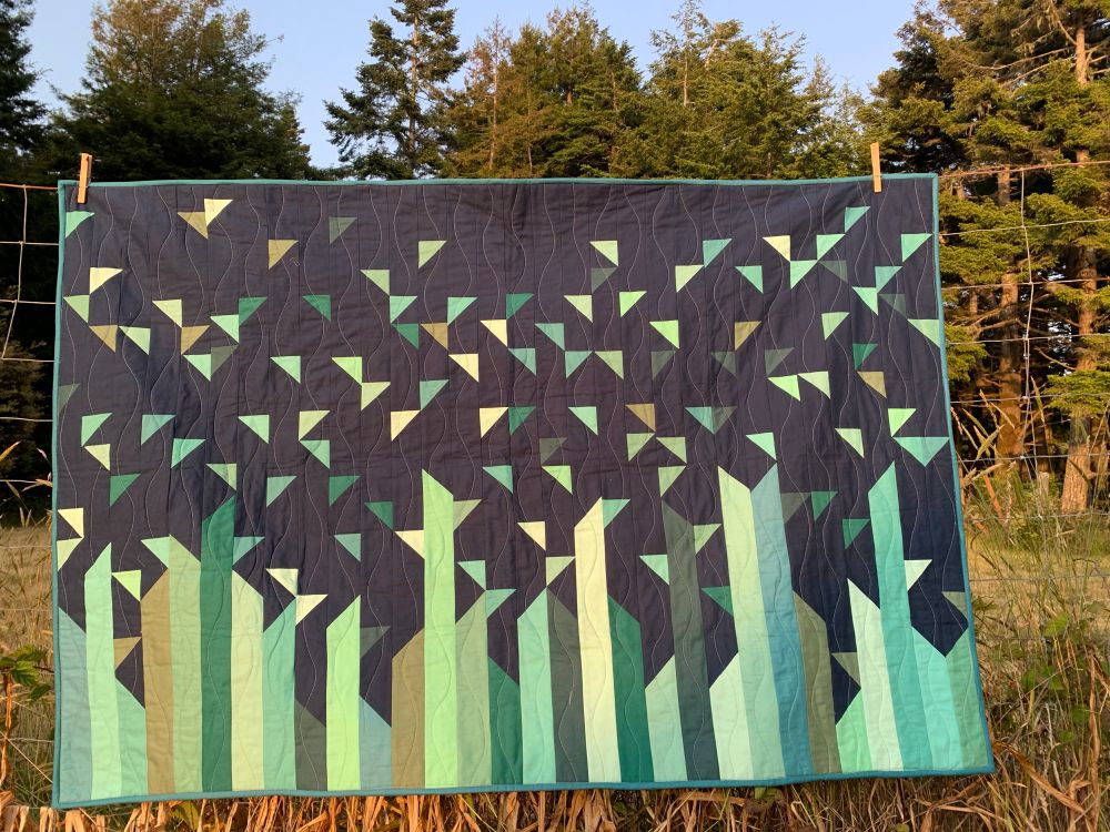 A strip-pieced quilt with random lengths in various shades of green melting into a scatter of half-square triangles against a dark blue background. 