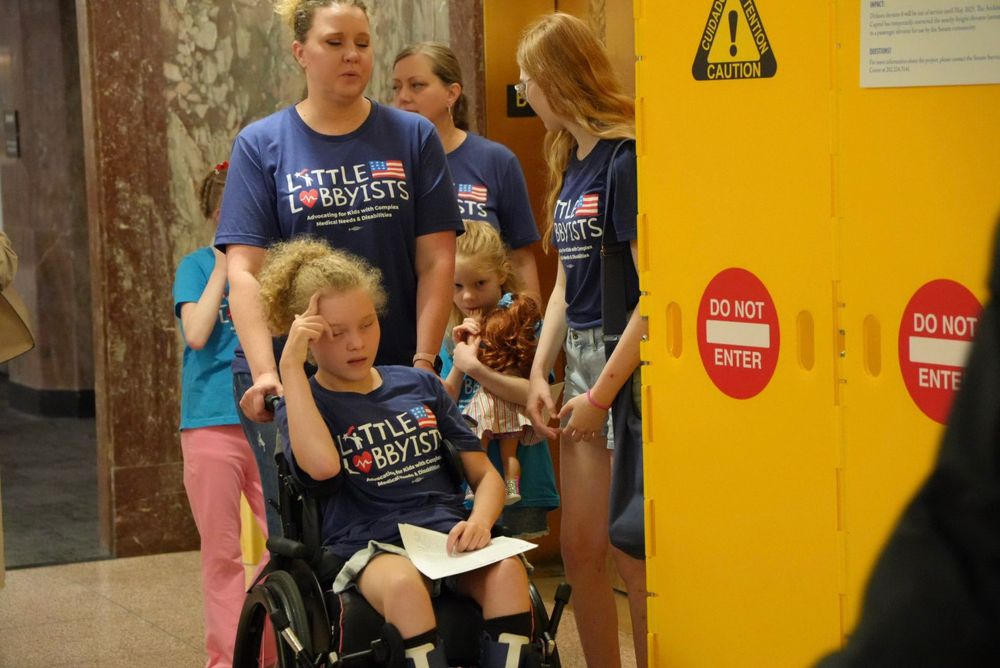 A blond girl in a wheelchair being pushed by her mother past a "do not enter" barrier. Several other children and parents are behind them. All wear shirts identifying them as members of Little Lobbyists.
