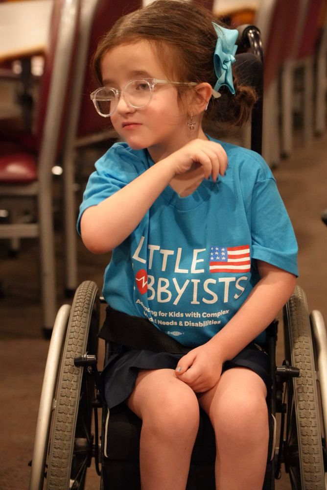 A brunette girl with glasses sitting in her wheelchair, face turned slightly away to look at someone off camera.