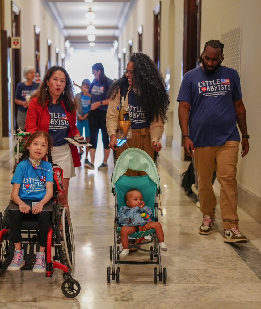 An Asian American woman pushing her daughter in a wheelchair, alongside a Black woman pushing her son in a stroller, accompanied by her husband. All are wearing "Little Lobbyists" shirts and the women are in the middle of a friendly exchange.