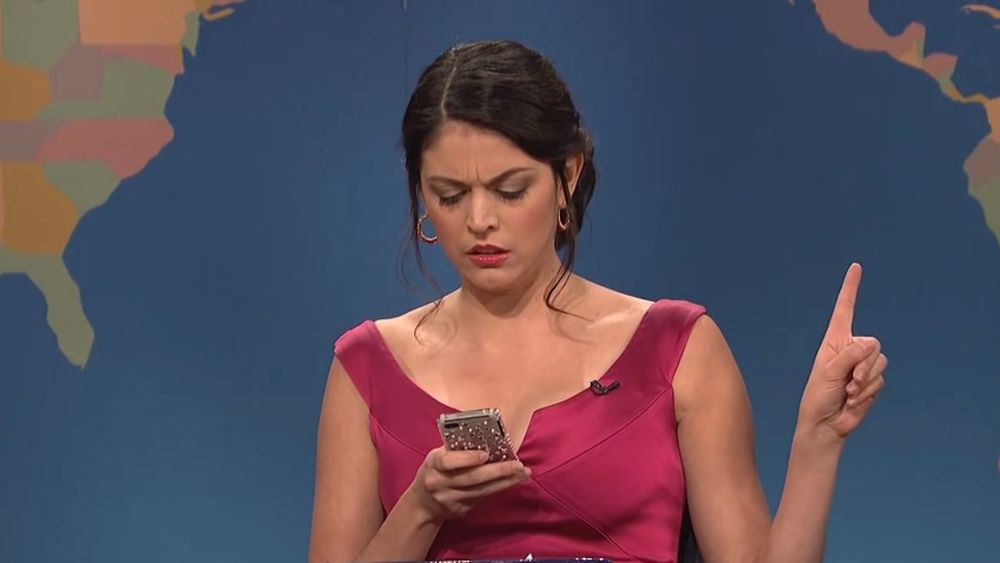 Cecily Strong on the set of SNL's Weekend Update in a pink sleeveless top looking at a smartphone with a "wait a minute" finger up, in character as the Girl You Wish You Hadn't Gotten Into a Conversation With at a Party