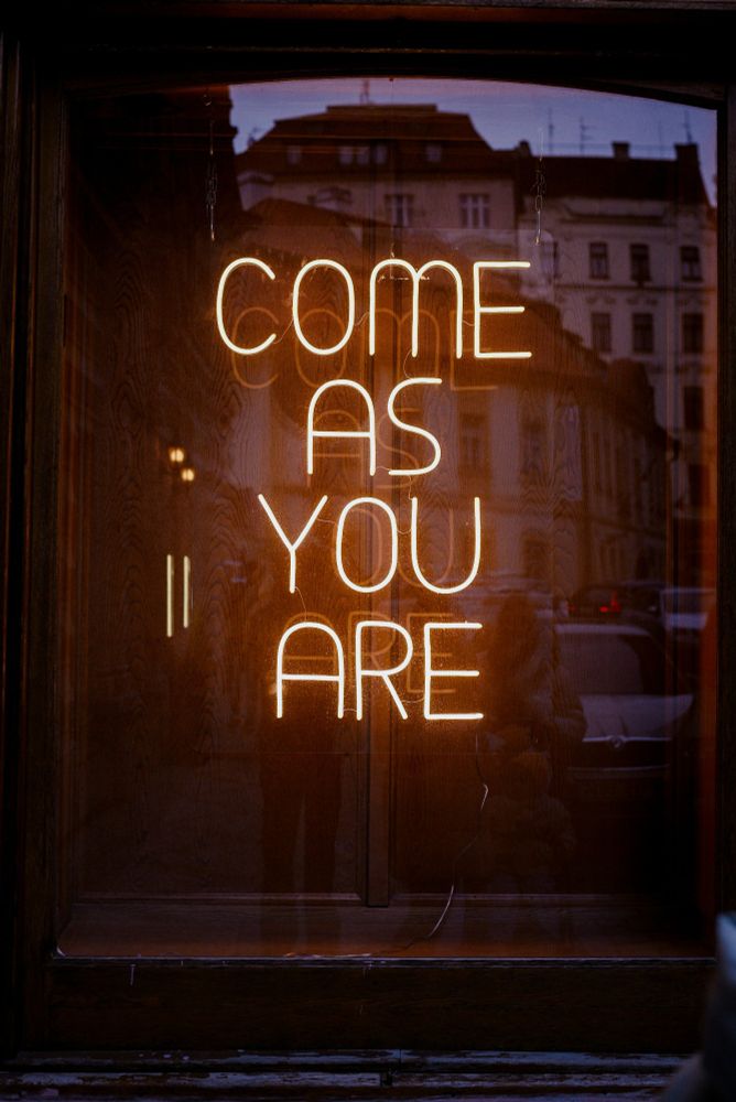 Illuminated sign reading "come as you are" with a building reflected in the background; photo by Kate Tepl on Unsplash